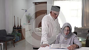 Muslim senior man teaching wife reading Koran or Quran in living room