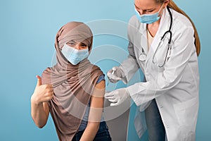 Muslim teenage girl in hijab and medical mask getting covid vaccine injection from female doctor, gesturing thumb up