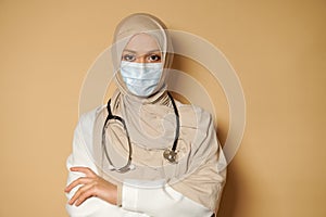 Muslim woman doctor in hijab and medical mask looking at camera with crossed arms. Beige background with copy space