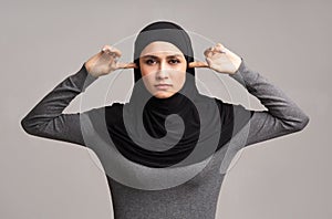 Muslim woman in hijab closing ears with fingers over grey background