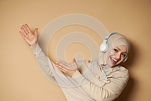 Muslim woman in hijab listening to the music in headphones, pointing hands on beige background with copy space