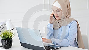 Muslim woman in hijab is talking mobile phone and typing on laptop at office work.