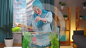 Muslim woman in national headscarf studies a book on aquariums and feeds goldfish in an aquarium