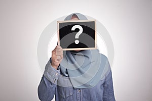 Muslim woman wearing hijab covering her face with blackboard written question mark on it, anonymouse facial expression