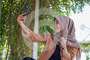 A Muslim woman wearing a hijab is taking a selfie using a smartphone with a smiling pose and her hands on her chest