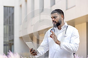 Muslim young male doctor standing on the street near the hospital and talking on a call from a tablet, online