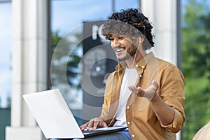 Muslim young man sitting on a bench on a city street wearing headphones and smilingly talking on a video call on a photo