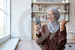 Muslim young woman at home in kitchen standing and using mobile phone, happy showing victory gesture with hand, reading