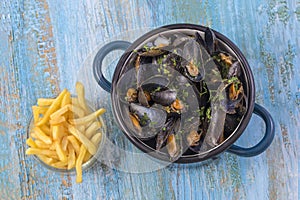 Mussels in a blue ceramic pot on a blue wooden background. with a glass bowl of french fries. Meditteranean lifestyle.