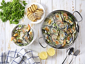 Mussels clams in cream sauce in cooking pan and toasted baguet on wooden table, top view