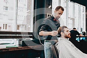 Mustachioed barber dressed in a black shirt with a red bow tie makes a stylish hairstyle to young man in a barbershop