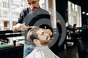 Mustachioed barber dressed in a black shirt with a red bow tie makes a stylish hairstyle to young man in a barbershop