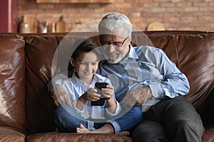 Happy little grandkid sit on sofa use cellphone with grandfather