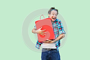 It is my! Crazy happy young adult shopoholic man in white t-shirt and checkered shirt standing, hugging shopping bags with toothy