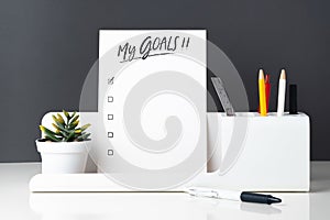 My goals list on notepad at modern office stationery on white table and dark grey wall.pencil, notepad,ruler,pen and pencil box