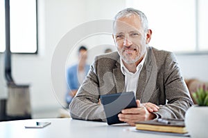 My new business partner. Portrait of a mature businessman using a digital tablet while sitting at a table in an office.