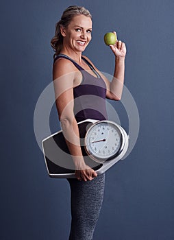 This is my secret. Studio portrait of an attractive mature woman holding an apple and a weightscale against a blue