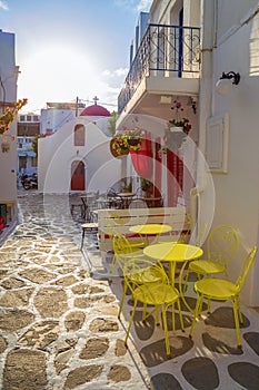 Mykonos streetview at sunrise with chapel and yellow chairs and tables, Greece