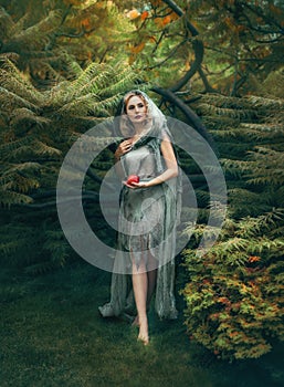 Mysterious evil witch with blond curly hair comes out of a thick forest with a red apple, in an old linen dress that