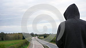 A mysterious hooded figure, back to camera. Looking down on an empty motorway in the countryside. With copy space