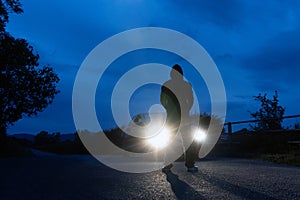 A mysterious hooded figure, silhouetted against the headlights of a parked car. On a lonely spooky country road at night