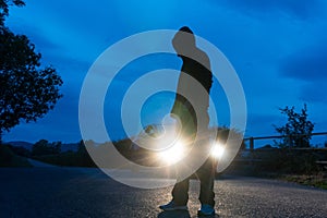 A mysterious hooded man, back to camera. looking at a long straight road, Back lighted by car headlights. On a summers evening. UK