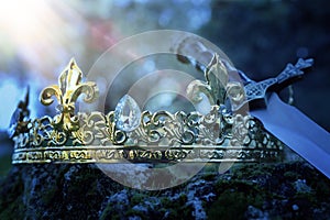 mysterious and magical photo of silver king crown and sword over the stone covered with moss in the England woods or field