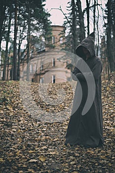 Mysterious monk, wizard or adept of secret society in hooded robe stands in forest against the background of medieval castle or