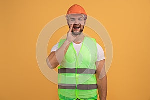 Mysterious surprised labourer worker looks with bugged eyes, whispers something, keeps palm near opened mouth, wears hardhat and