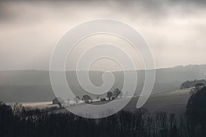 Mysterious winter foggy landscape with hills, trees covered in frost against a backdrop of dense fog by the early morning sun.