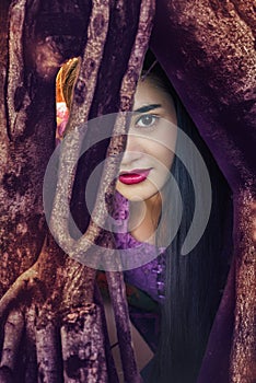 Mysterious woman,Beautiful woman with long dark hair and red lips resting in the tree roots and looking at you.