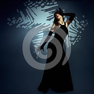 Mysterious Woman in long Black Dress perform on Art Stage. Lady in evening Gown over Shadow Background. Beautiful Girl dancing