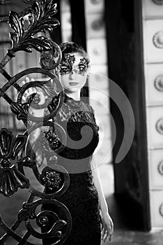 Mysterious woman in venetian carnival mask near wrought iron gate. Noir style