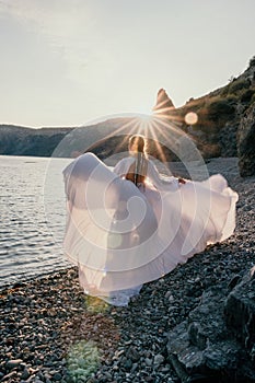 Mysterious young woman with braids in long white dress alone on the beach. Sunset over the sea with rocky volcanic cliff