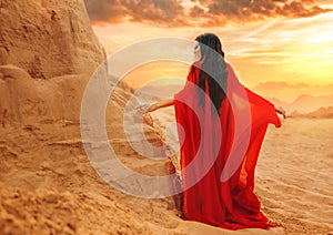 Mystery arabic woman in red long dress stands in desert long train silk fabric fly flutter in wind motion. clothes gold