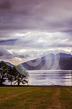 Mystic landscape lake scenery in Scotland: Cloudy sky, meadow, trees and lake with sunbeams, mountain range in the background.