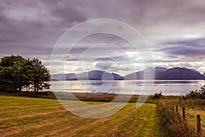Mystic landscape lake scenery in Scotland: Cloudy sky, meadow, trees and lake with sunbeams, mountain range in the background.