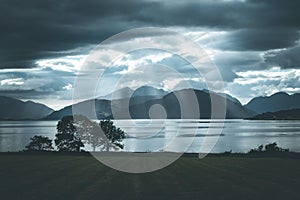 Mystic landscape lake scenery in Scotland: Cloudy sky, meadow, trees and lake with sunbeams, mountain range in the background.