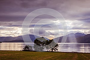 Mystic landscape lake scenery in Scotland: Cloudy sky, meadow, trees and lake with sunbeams, mountain range in the background.