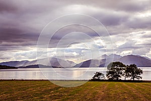 Mystic landscape lake scenery in Scotland: Cloudy sky, meadow, trees and lake with sunbeams, mountain range in the background.