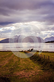 Mystic landscape lake scenery in Scotland: Cloudy sky, meadow, trees and lake with sunbeams, mountain range in the background.