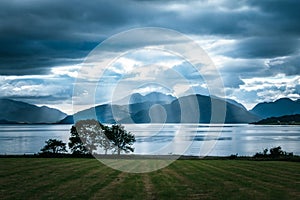 Mystic landscape lake scenery in Scotland: Cloudy sky, meadow, trees and lake with sunbeams, mountain range in the background.