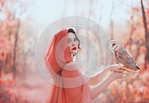 Mystical pagan woman with covered head in peach scarf in forest, holds barn ow