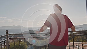 A n man stands on a balcony overlooking the sea and mountains on a sunny day.