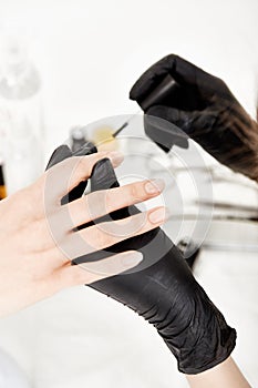 Nail artist in gloves holding woman hand applying clear polish.