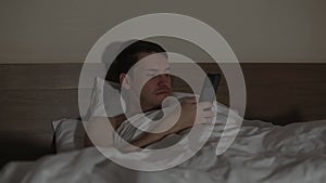 Naked man lying on pillow under blanket in bed with mobile phone in late evening. Relaxed male browsing smartphone at