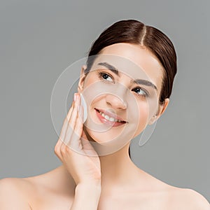 Naked woman cleaning face with cotton pad and smiling isolated on grey