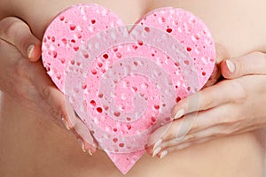 Naked woman holding pink heart sponge in hands.