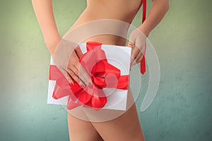 Naked woman holds a gift box with red ribbon next to her hips against colorful textured background.
