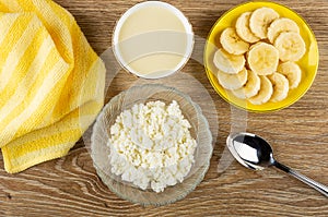 Napkin, bowl with condensed milk, cottage cheese in bowl, saucer with slices of banana, spoon on wooden table. Top view